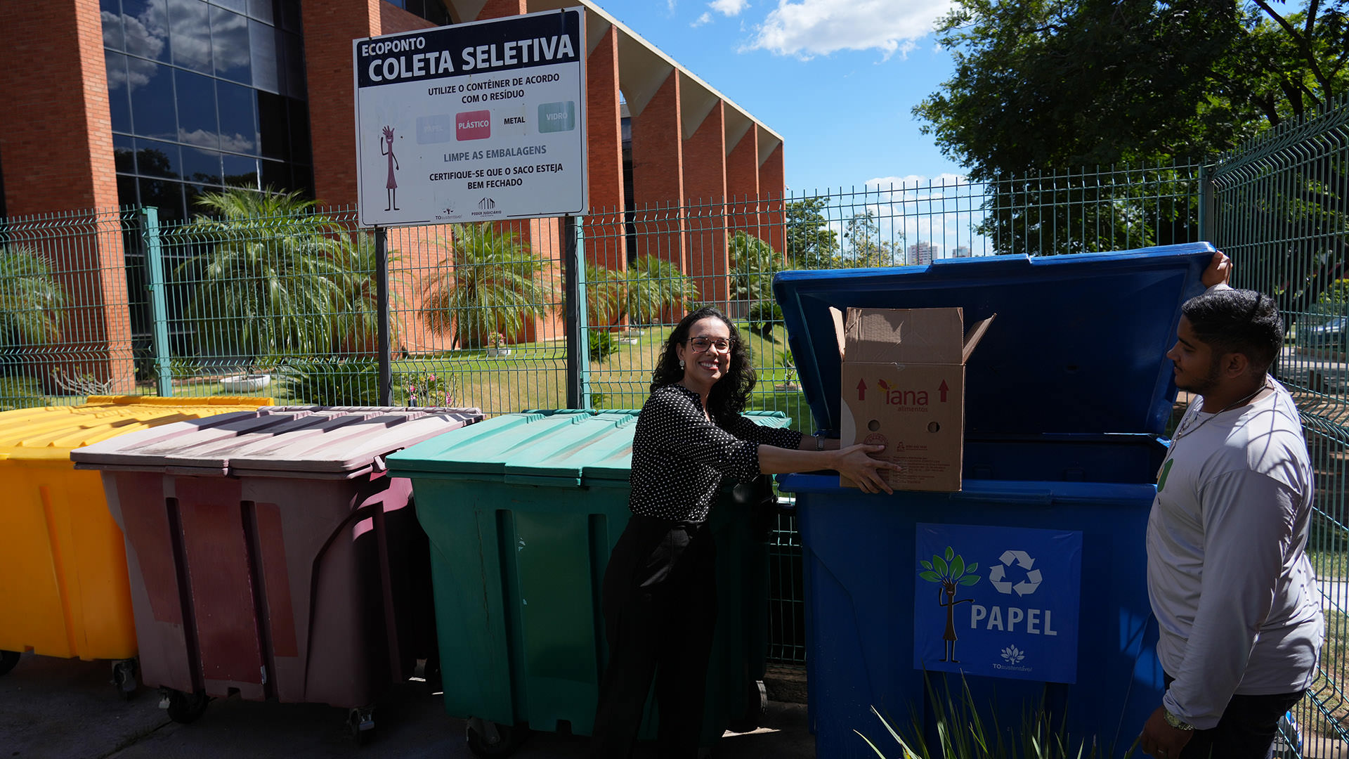 A imagem mostra uma servidora realizando o descarte de materiais recicláveis no ecoponto de coleta seletiva instalado no Tribunal de Justiça do Tocantins (TJTO), durante o Dia D da Coleta Seletiva Solidária, enquanto um colaborador auxilia na abertura do contêiner.