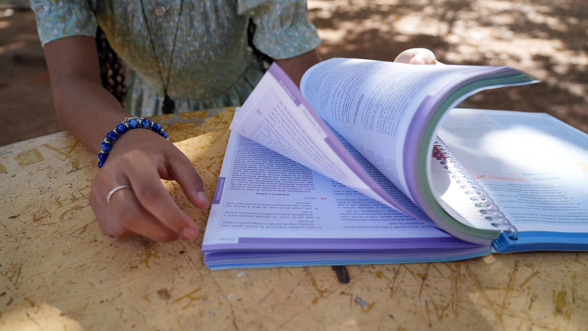 A foto mostra uma adolescente folheando um livro didático enquanto estuda em uma área externa. O registro destaca apenas parte do corpo da jovem, preservando sua identidade. Sobre a mesa amarela, os livros abertos reforçam o ambiente de aprendizado e dedicação aos estudos.
