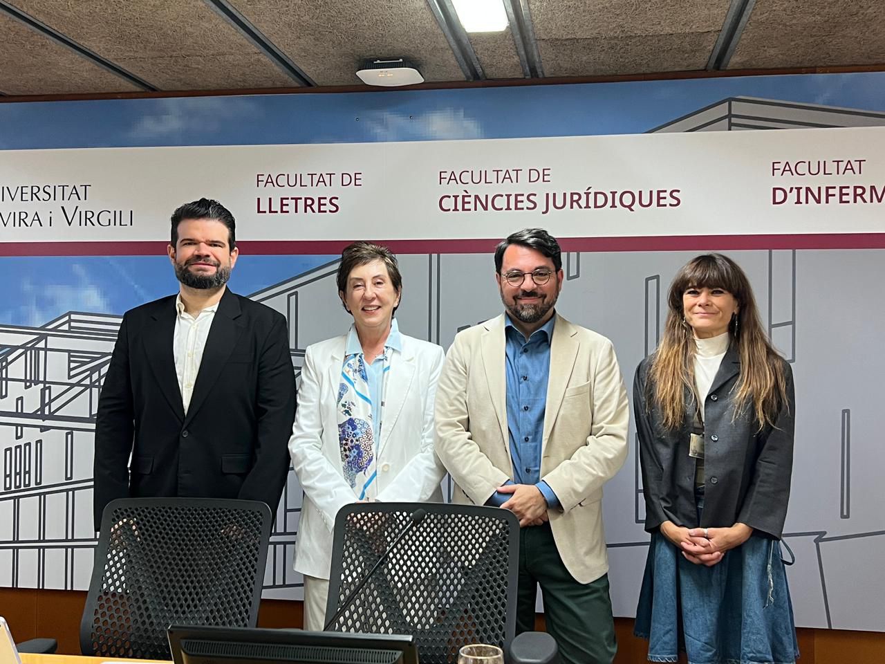 A imagem mostra quatro pessoas em pé, posando para uma foto, em frente aos painéis das faculdades da Universitat Rovira i Virgili.  O grupo aparece sorrindo e vestido com trajes sociais.