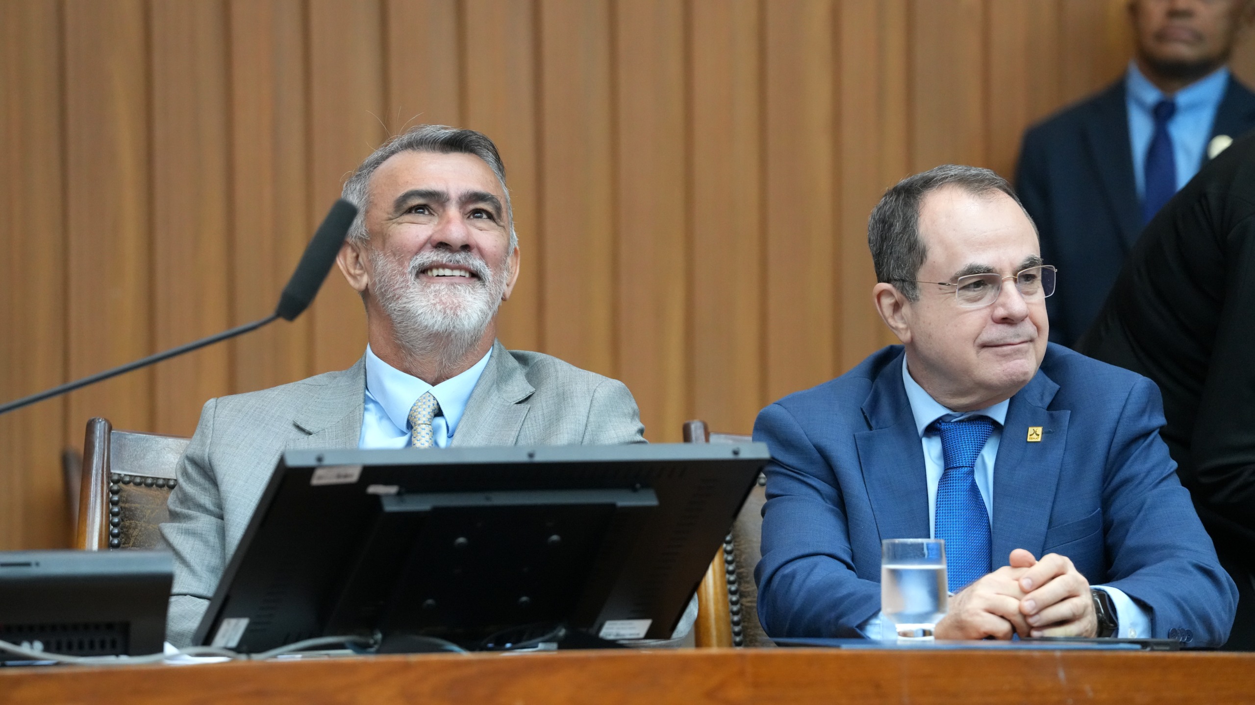A imagem mostra o presidente da Assembleia Legislativa, deputado Amélio Cayres, e o desembargador Marco Anthony Steveson Villas Boas, diretor-geral da Esmat, sentados à mesa de solenidade em um ambiente institucional. Vestidos de terno, os dois acompanham atentamente o evento, tendo ao fundo um painel de madeira que compõe o cenário oficial.
