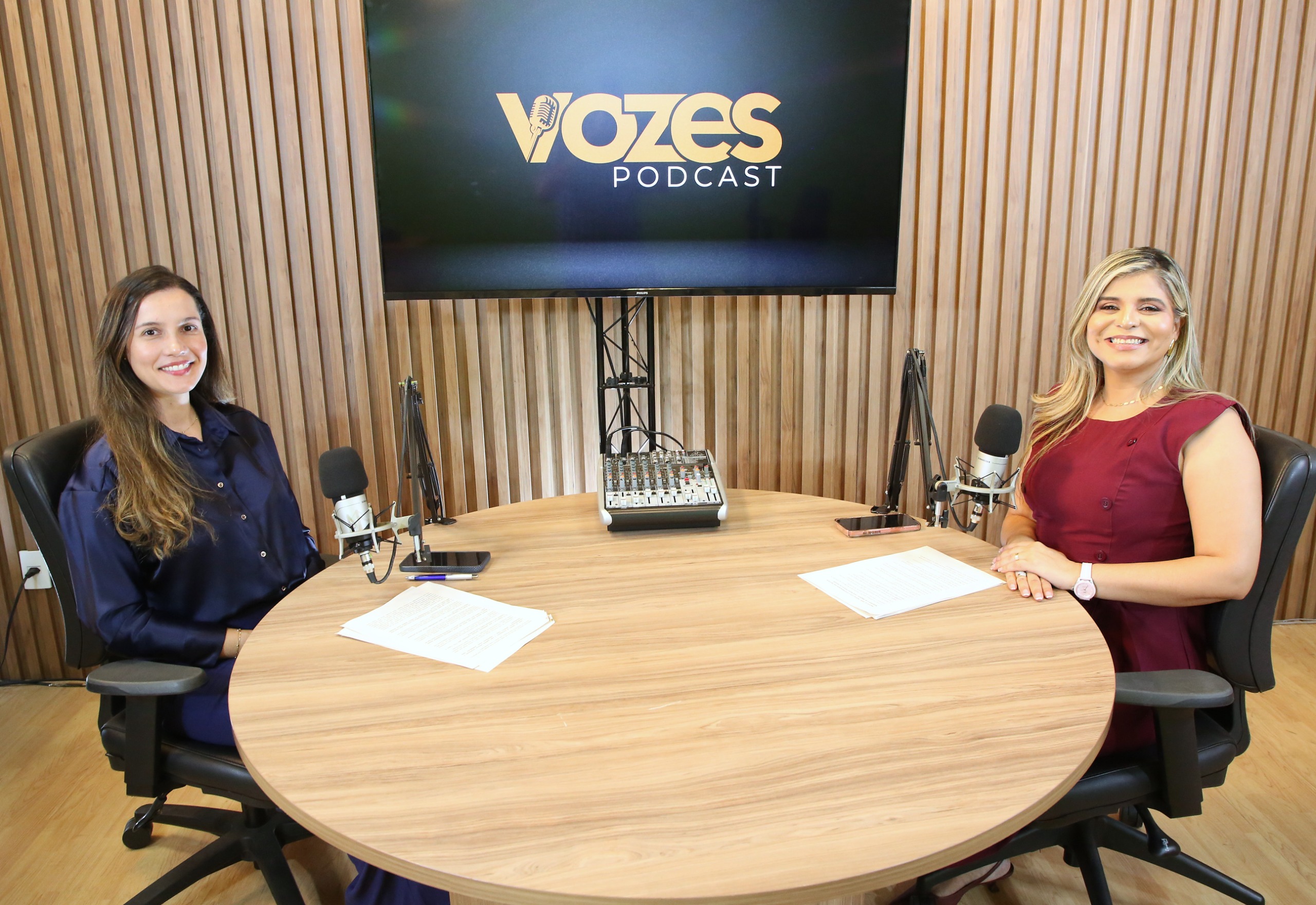 A imagem mostra duas apresentadoras do Podcast Vozes durante a gravação da 13ª edição do programa, em estúdio equipado com microfones e mesa de som, tendo ao fundo a identidade visual do podcast.