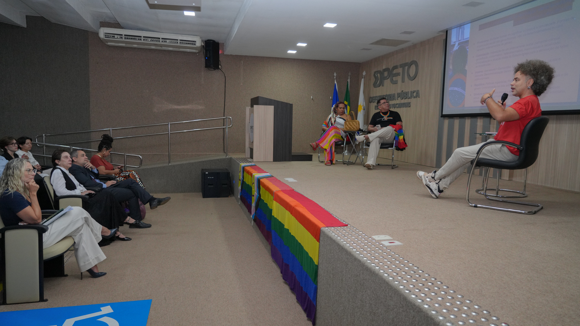 A imagem mostra um painel de debates do workshop “Nenhum Direito a Menos: Democracia e Cidadania”, promovido pela Defensoria Pública do Tocantins, reunindo especialistas e representantes de instituições em discussão sobre direitos da população LGBTQIAPN+. No auditório, autoridades e participantes acompanham atentamente as exposições.