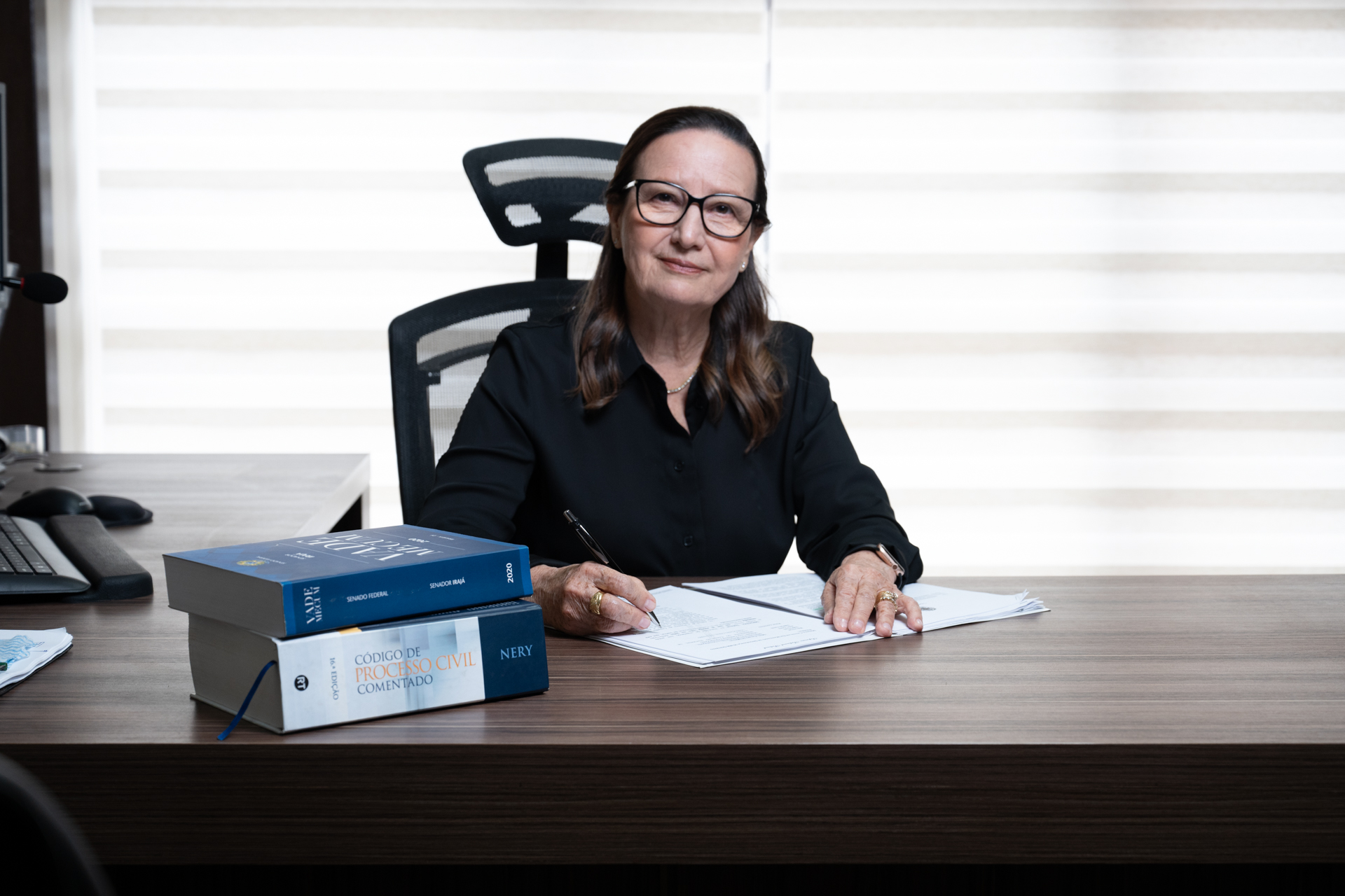 A presidente do TJTO, desembargadora Jacqueline Adorno, assina documentos em seu gabinete, ao lado de livros jurídicos sobre a mesa.