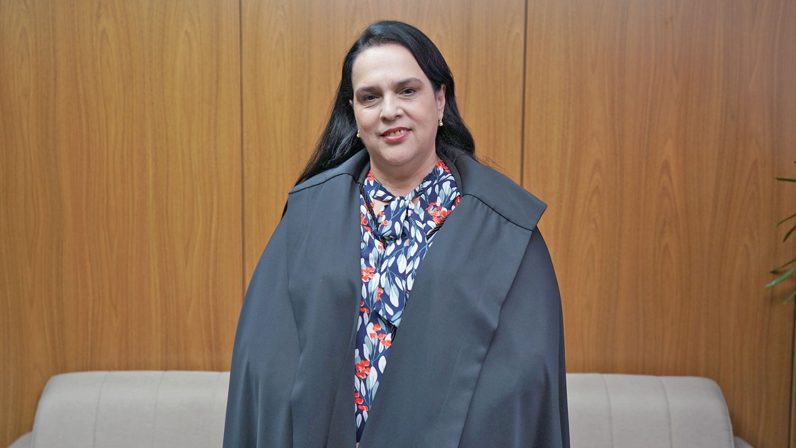 Desembargadora Jacqueline Adorno posa para fotografia institucional nas dependências do Tribunal de Justiça do Tocantins. Vestida com a toga da magistratura, ela aparece em pé, diante de painel amadeirado, com expressão serena e olhar direcionado à câmera. Ao fundo, há um sofá em tom claro, compondo o ambiente interno.