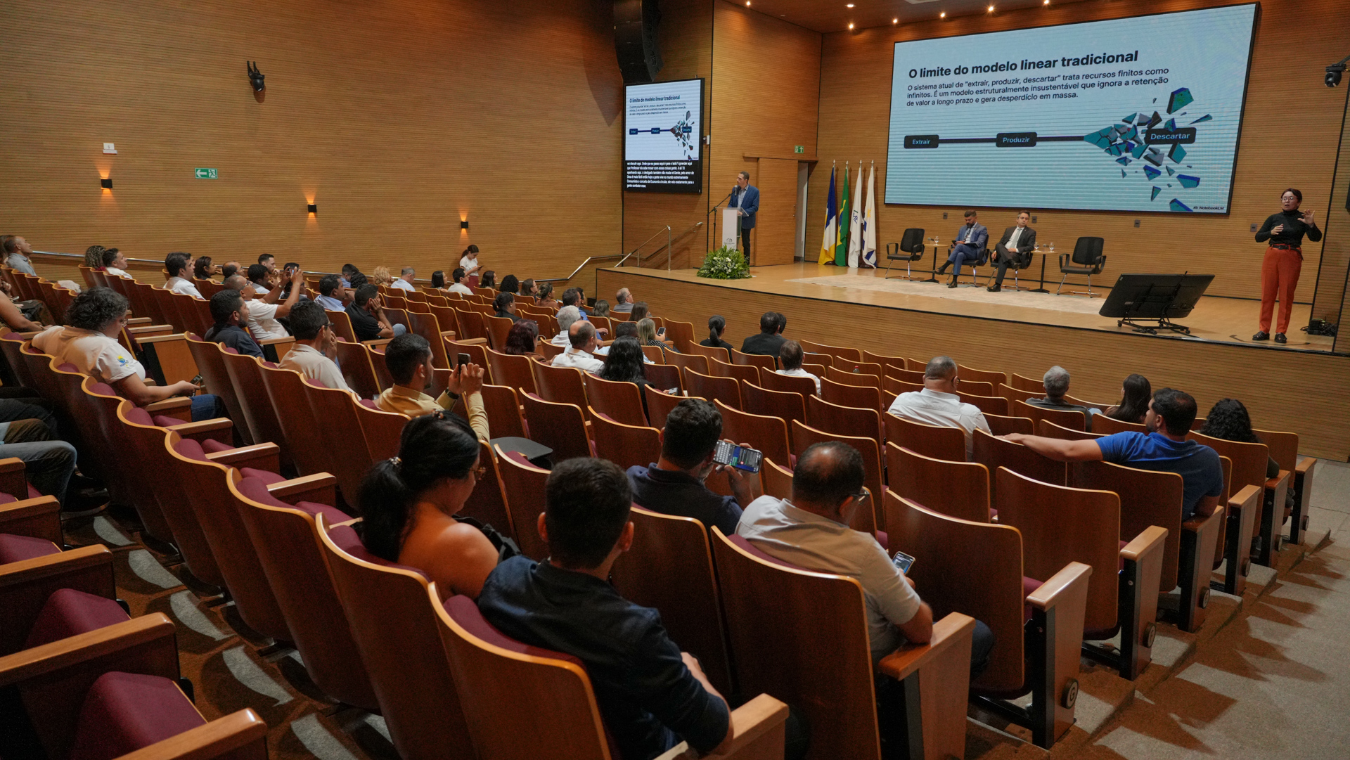 A imagem mostra o auditório do Tribunal de Justiça do Tocantins (TJTO) durante a programação da tarde do Seminário de Resíduos Sólidos, com participantes acompanhando a palestra sobre economia circular e gestão sustentável dos resíduos.  No palco, os mediadores e palestrantes conduzem os debates, enquanto uma intérprete de Libras garante a acessibilidade do evento promovido no âmbito da II Semana da Pauta Verde.