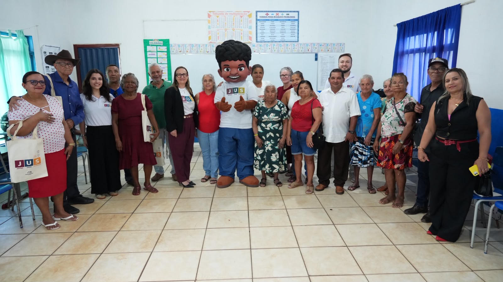 A presidente do TJTO visita a oficina voltada às pessoas idosas durante o projeto JUS em Ação, acompanhada do mascote Justino. Na sala de aula, ela posa ao lado dos participantes e da equipe organizadora, em um momento de integração, acolhimento e valorização da terceira idade.