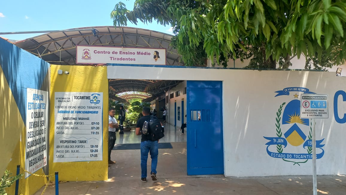 A imagem mostra a entrada do Centro de Ensino Médio Tiradentes, local de aplicação do Exame Nacional dos Cartórios (ENAC), com um candidato acessando o prédio para realizar a prova.