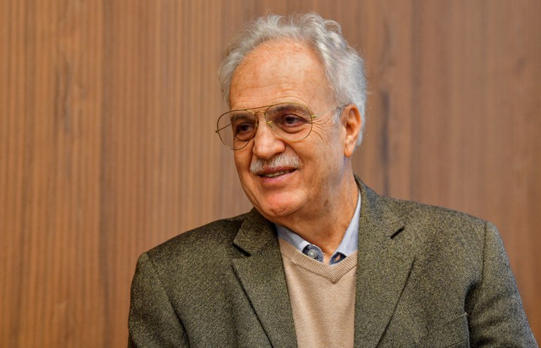 O climatologista Carlos Nobre aparece em plano médio, sentado, com expressão serena e leve sorriso. Ele veste blazer cinza, suéter bege sobre camisa azul-clara e óculos de armação metálica. Ao fundo, um painel de madeira em tons claros compõe o ambiente interno.