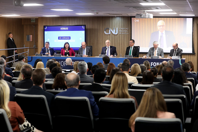 A imagem mostra a mesa de abertura de um evento institucional realizado no auditório do Conselho Nacional de Justiça (CNJ). À frente do auditório, seis autoridades compõem a mesa principal, posicionadas atrás de uma bancada identificada com a marca do Fórum Nacional do Judiciário para a Saúde (Fonajus). Ao centro, os participantes acompanham a solenidade, enquanto um dos palestrantes tem sua imagem projetada em um grande telão ao fundo.  O auditório está lotado, com dezenas de participantes sentados e voltados para a mesa de autoridades. No painel lateral, é possível visualizar a identificação da “VIII Jornada de Direito da Saúde”, reforçando o caráter técnico e jurídico do encontro. O ambiente apresenta estrutura moderna, com iluminação adequada e recursos audiovisuais utilizados para a transmissão e acompanhamento das atividades do evento.