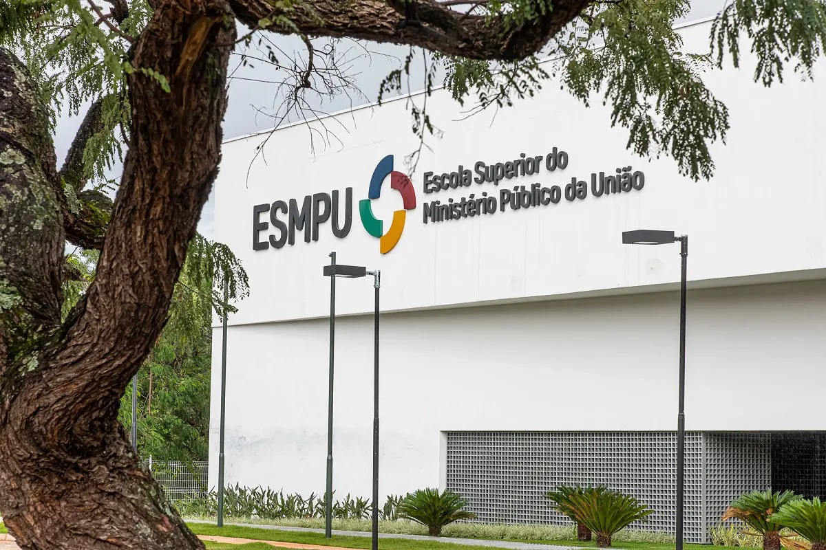 Fotografia da fachada da Escola Superior do Ministério Público da União (ESMPU). O prédio tem parede branca com a logomarca colorida da instituição e a inscrição “ESMPU – Escola Superior do Ministério Público da União”. Em primeiro plano, uma árvore de tronco grosso e galhos espalhados enquadra a imagem. À frente do edifício há postes de iluminação, gramado e plantas ornamentais. O céu está nublado.