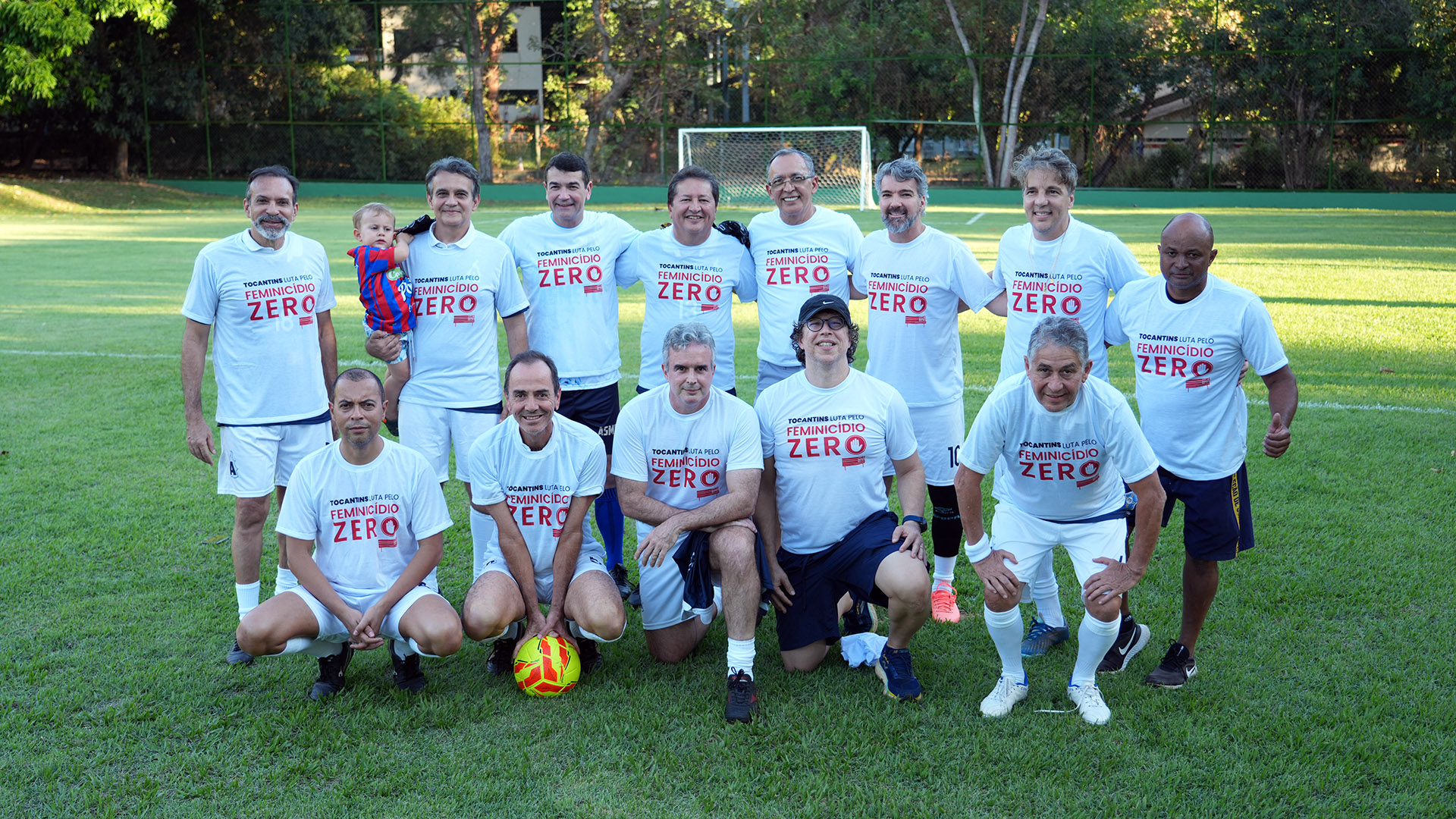 Magistrados participantes do Campeonato Nacional de Futebol Sênior da Magistratura posam para uma foto coletiva em campo, vestindo camisetas da campanha “Tocantins Luta pelo Feminicídio Zero”. O grupo está reunido no gramado antes da partida, em um gesto de apoio à conscientização e ao enfrentamento da violência contra a mulher. Ao fundo, aparecem as traves do campo e a área arborizada da sede campestre da Asmeto, em Palmas.