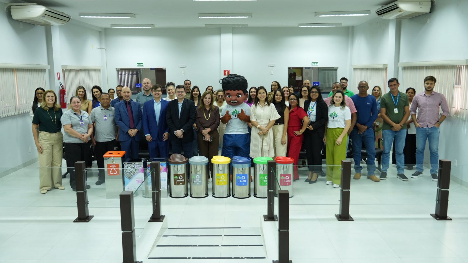 Fotografia de grupo registrada no hall da Comarca de Palmeirópolis. Ao centro, está o mascote Justino, personagem com camiseta branca e calça azul, cercado por cerca de 30 pessoas entre magistrados(as), servidores(as), colaboradores(as) e representantes de instituições parceiras. Na primeira fila, destaca-se a desembargadora Ângela Prudente ao lado dos participantes. À frente do grupo estão dispostos conjuntos de lixeiras da coleta seletiva identificadas por cores e categorias de resíduos, além de coletores para materiais específicos. O ambiente é amplo, iluminado e climatizado, reforçando o contexto de uma ação voltada à sustentabilidade e à educação ambiental.