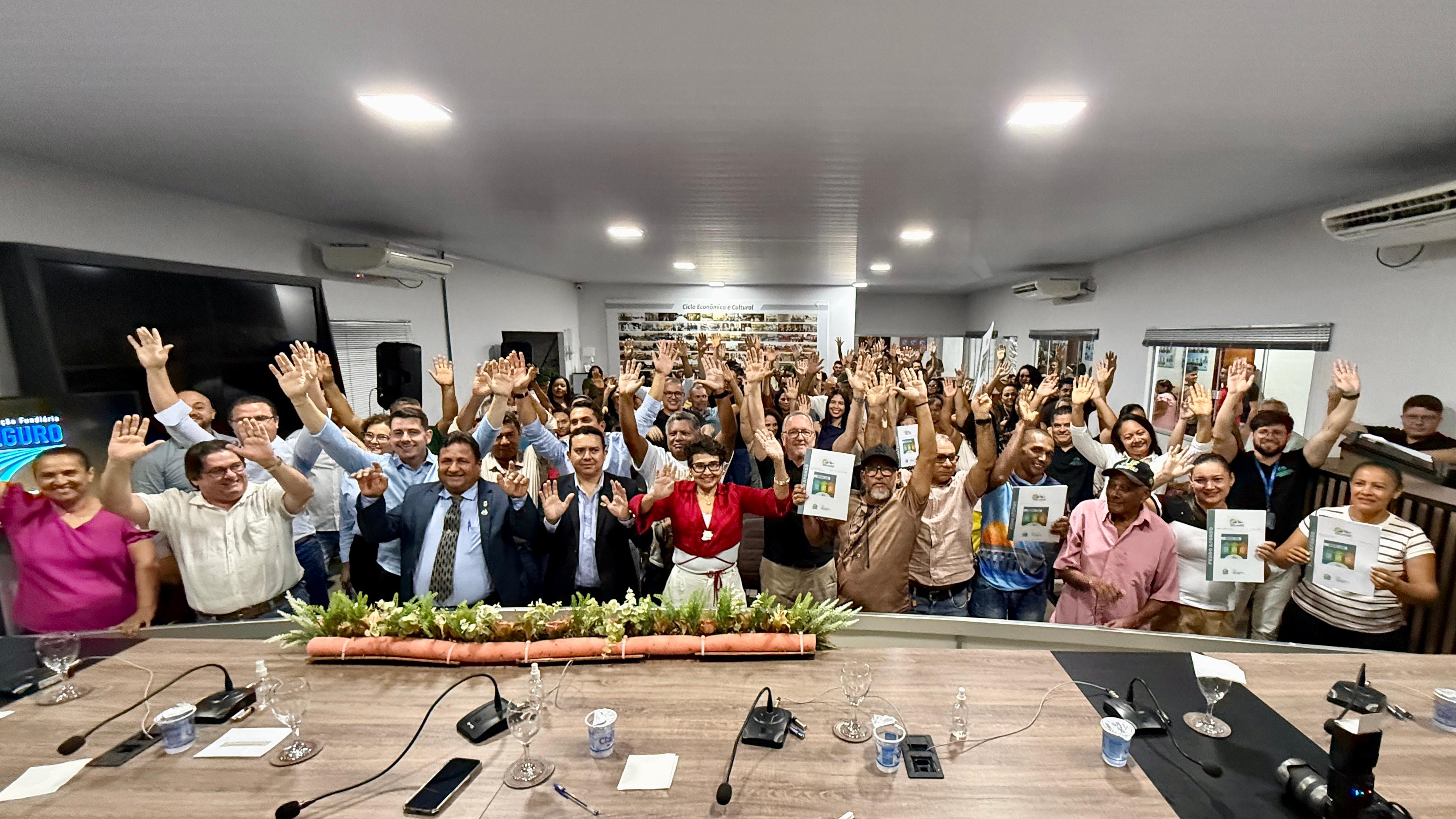 Fotografia feita em um auditório durante evento institucional. Em primeiro plano, atrás de uma mesa de reuniões equipada com microfones, copos e arranjo floral, dezenas de pessoas posam para a foto coletiva com os braços erguidos. No centro da imagem estão autoridades e participantes do encontro, enquanto o público ocupa todo o espaço do auditório ao fundo. Algumas pessoas seguram exemplares de um material impresso. O ambiente é iluminado, com telões, aparelhos de ar-condicionado e painéis nas paredes, transmitindo clima de participação, engajamento e celebração coletiva.