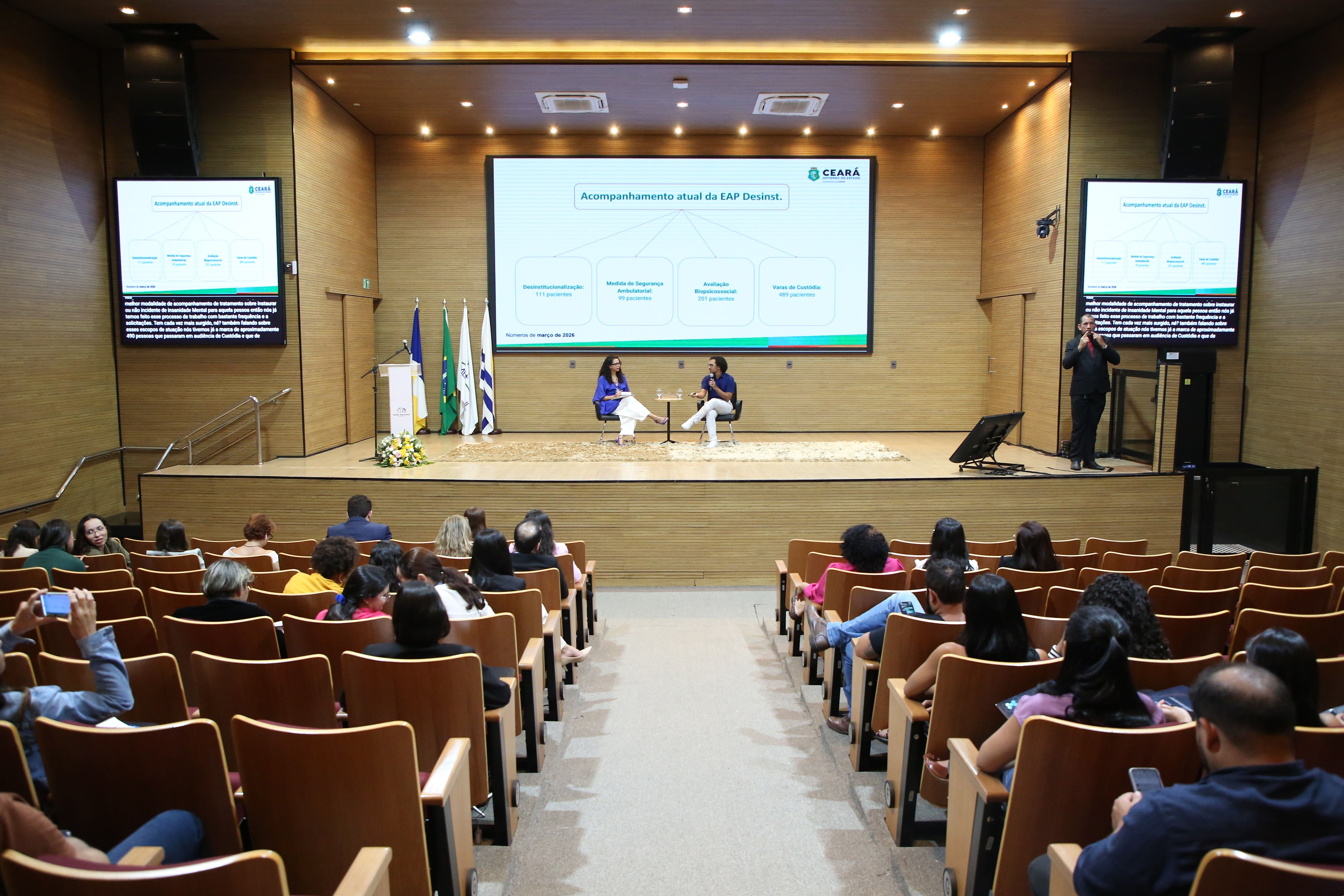 A imagem mostra o auditório com dezenas de participantes acomodados em poltronas, acompanhando as atividades do Simpósio sobre a Política Antimanicomial no Tocantins. No palco, dois palestrantes participam de uma mesa de diálogo, enquanto a apresentação é exibida em telões e conta com tradução em Libras.