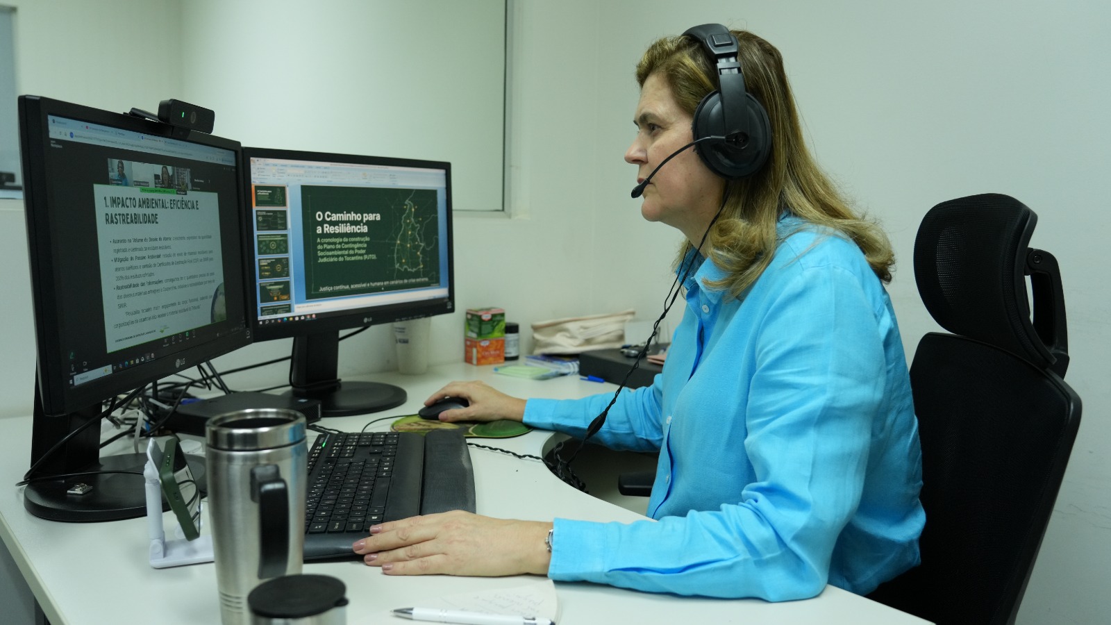 Luciene Dantas participa da videoconferência em seu ambiente de trabalho. Sentada à mesa, ela usa fone de ouvido com microfone e opera o computador enquanto acompanha uma apresentação exibida em dois monitores. Em uma das telas, há um slide sobre impacto ambiental, eficiência e rastreabilidade; na outra, uma apresentação intitulada "O Caminho para a Resiliência", relacionada ao Plano de Contingência Socioambiental do Poder Judiciário do Tocantins. Sobre a mesa estão teclado, mouse, um copo térmico, bloco de anotações, caneta e materiais de escritório. O ambiente é um escritório com mobiliário corporativo.