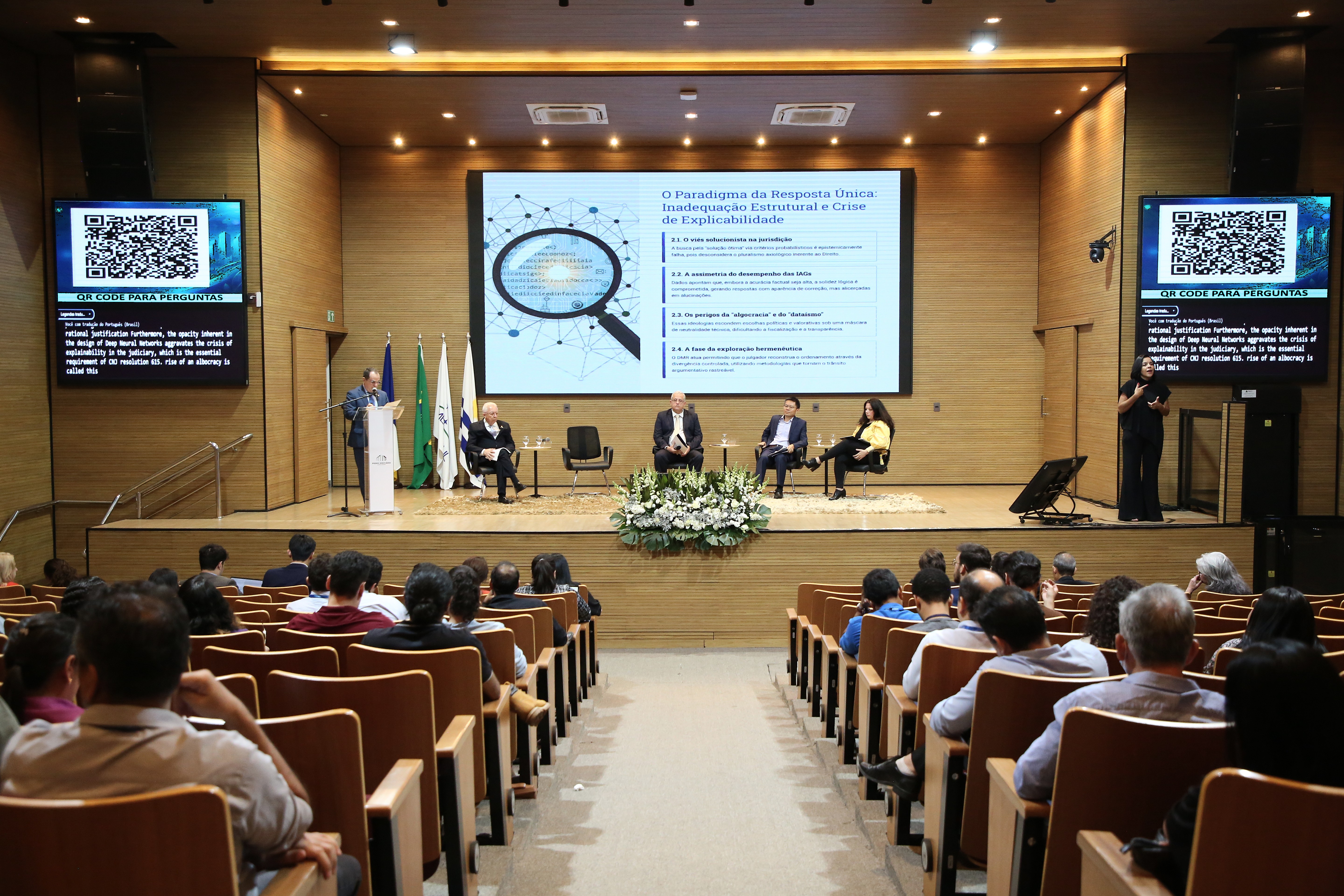 Participantes acompanham palestra durante evento realizado em auditório, com mesa de debates, apresentação em telão e tradução simultânea em Libras.