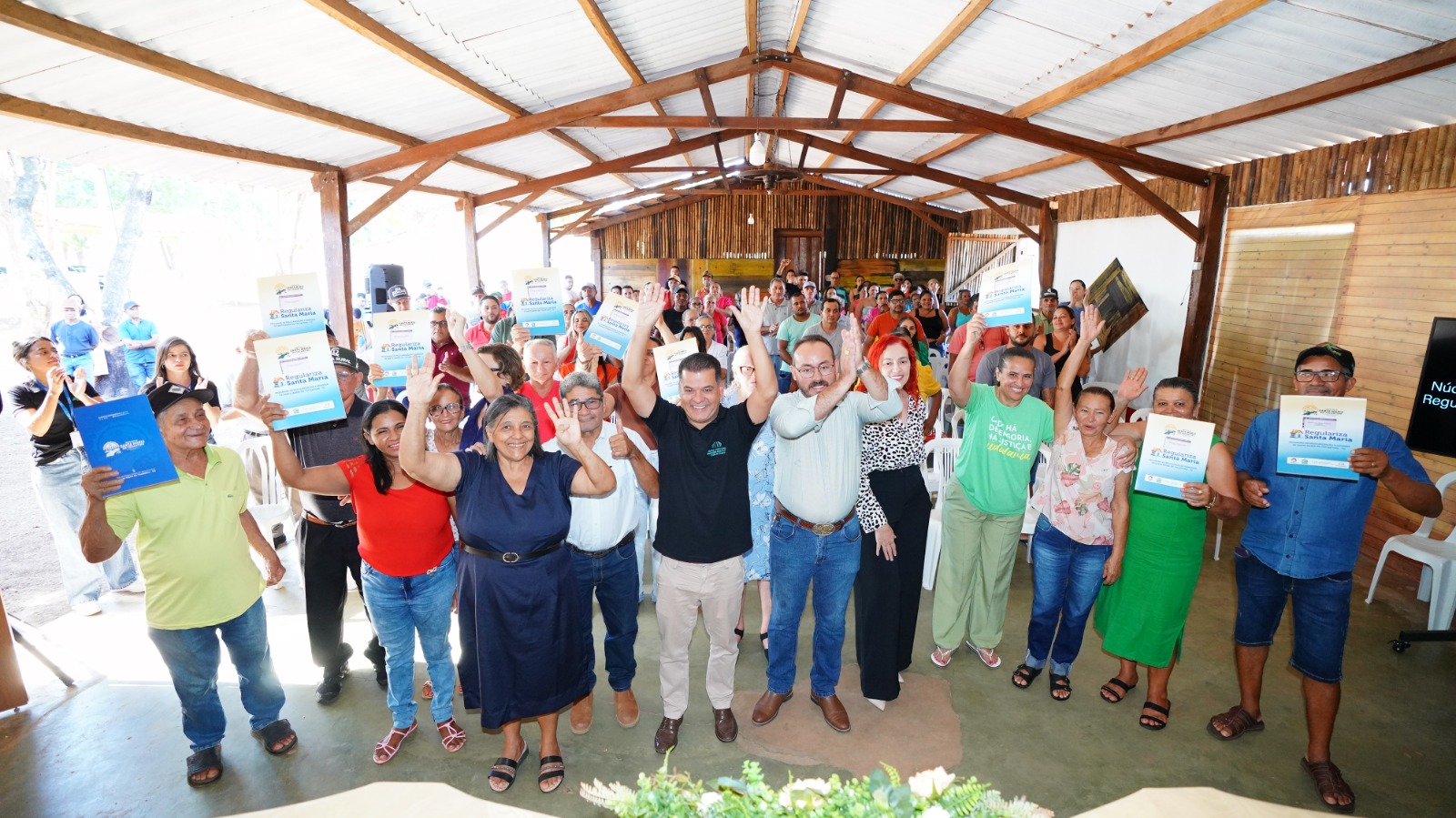 A imagem mostra moradores de Itacajá reunidos em um espaço comunitário durante a entrega de títulos de regularização fundiária, celebrando a conquista do direito à propriedade. Em clima de comemoração, participantes exibem os documentos recebidos ao lado de autoridades e representantes do projeto, marcando um momento histórico para quase 200 famílias de Santa Maria e Itacajá.