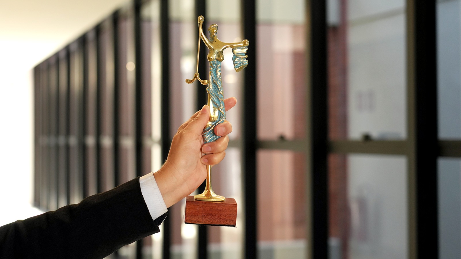 Uma mão segura um troféu dourado em formato da deusa da Justiça, com a balança em destaque, simbolizando equilíbrio, ética e imparcialidade. Ao fundo, desfocado, aparece um corredor com amplas janelas de vidro, remetendo ao ambiente institucional do Poder Judiciário.
