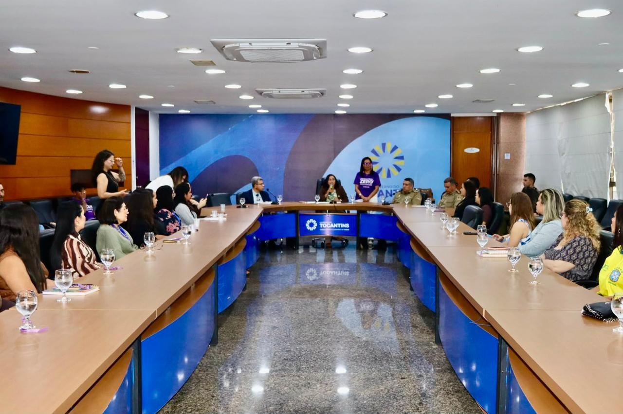 Imagem mostra uma reunião com autoridades e representantes de instituições sentados à mesa em formato de "U", acompanhando uma apresentação sobre combate ao feminicídio