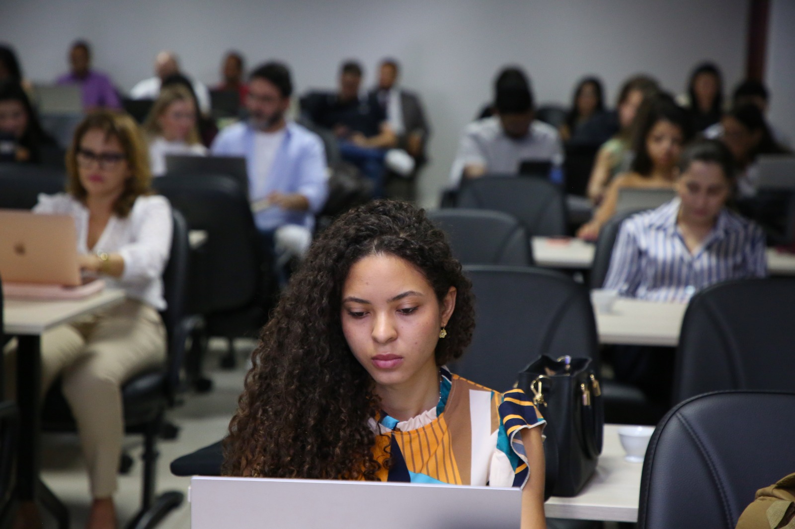 A imagem mostra uma sala de aula com diversos participantes acompanhando um curso em computadores, enquanto uma jovem em primeiro plano concentra-se na atividade em seu notebook.