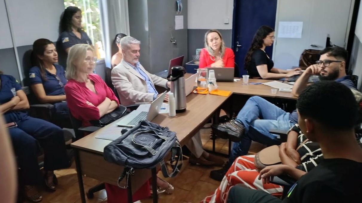 Em uma sala de reuniões, cerca de dez pessoas estão sentadas ao redor de uma mesa retangular durante uma audiência concentrada. Entre os participantes, há magistrados, servidores e representantes da rede de proteção, alguns utilizando notebooks. Ao centro da mesa há uma garrafa térmica, copos e materiais de trabalho. O ambiente é iluminado por luz natural que entra por uma janela à esquerda.