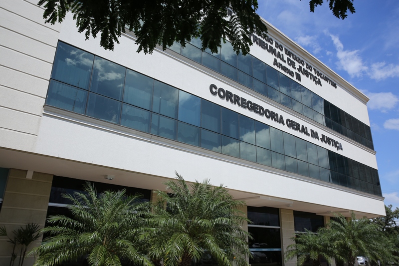 Foto externa do prédio da Corregedoria-Geral da Justiça do Tribunal de Justiça do Estado do Tocantins. A imagem mostra a fachada moderna do edifício, com revestimento em vidro espelhado e estrutura nas cores branca e bege. Em destaque, aparecem as inscrições “Corregedoria-Geral da Justiça” e “Tribunal de Justiça do Estado do Tocantins”. À frente do prédio, há jardins com palmeiras e vegetação ornamental. O céu azul com nuvens refletido nos vidros complementa a composição da fotografia.