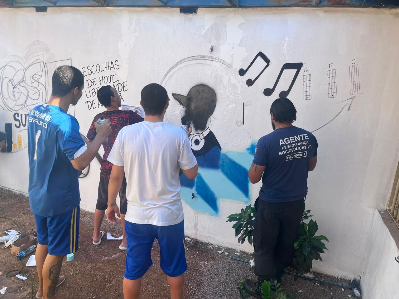 Adolescentes e agente socioeducativo participam da pintura de um mural artístico em área externa de unidade socioeducativa, durante atividade de grafite voltada à expressão cultural e ao protagonismo juvenil.