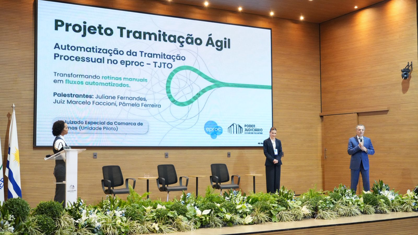 A foto mostra os palestrantes do painel “Projeto Tramitação Ágil – Automatização da Tramitação Processual no eproc – TJTO” durante apresentação no auditório do Tribunal de Justiça do Tocantins, no IV Encontro Interinstitucional do eproc. No palco, o juiz Marcelo Faccioni faz uso da palavra enquanto Juliane Fernandes e Pâmela Ferreira acompanham a exposição. Ao fundo, um telão exibe o tema da palestra e informações sobre o projeto desenvolvido no 1º Juizado Especial da Comarca de Palmas, unidade-piloto da iniciativa.