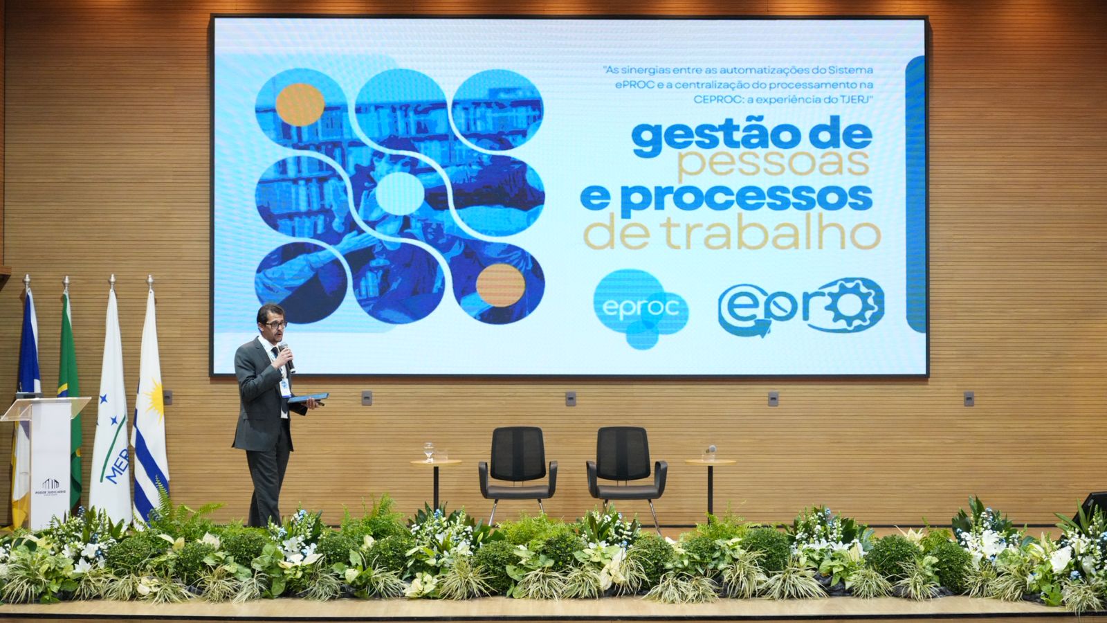 A imagem mostra o juiz Josué de Matos Ferreira, do Tribunal de Justiça do Estado do Rio de Janeiro (TJERJ), apresentando a palestra "As sinergias entre as automatizações do Sistema ePROC e a centralização do processamento na CEPROC: a experiência do TJERJ" no palco do auditório do TJTO.  Ao fundo, um telão exibe o tema da apresentação, com destaque para os conceitos de gestão de pessoas e processos de trabalho, enquanto o palestrante conduz a exposição aos participantes do IV Encontro Interinstitucional do eproc.