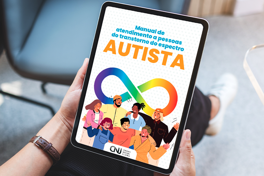 A imagem mostra uma pessoa segurando um tablet com as duas mãos, enquanto está sentada em um ambiente interno. Na tela do dispositivo aparece a capa do Manual de atendimento a pessoas do transtorno do espectro autista. O título está em letras azuis e laranja sobre um fundo branco. Ao centro, há o símbolo do infinito nas cores do arco-íris, associado à conscientização sobre o transtorno do espectro autista.