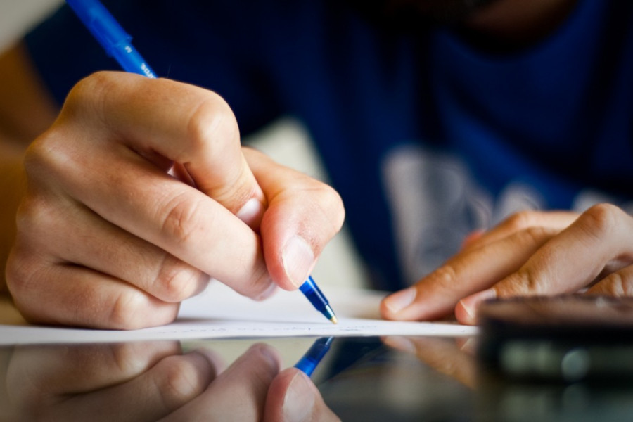 Close nas mãos de uma pessoa escrevendo com uma caneta azul sobre uma folha de papel. Em primeiro plano, há o reflexo da cena em uma superfície brilhante e parte de um celular desfocado. O foco da imagem está na escrita, remetendo à realização de prova, preenchimento de formulário ou produção de texto.