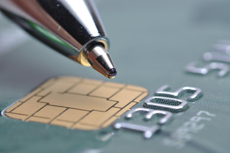 Imagem em close de um cartão bancário com chip dourado. Sobre o cartão, uma caneta metálica toca a superfície próxima ao chip, com foco na ponta da caneta e no chip. Ao fundo, desfocado, aparecem parte dos números em relevo do cartão. A imagem remete a pagamentos, serviços bancários e transações financeiras.