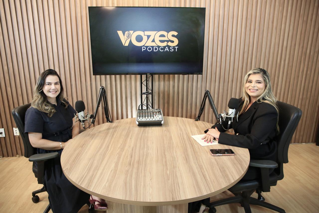 A imagem mostra duas mulheres sentadas em uma mesa redonda com microfones. As duas estão participando da gravação de um podcast.