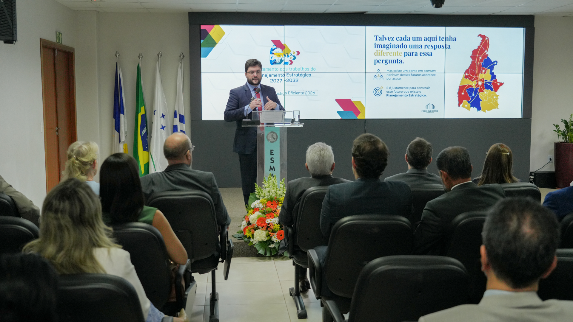 O coordenador da Coordenadoria de Gestão Estratégica, Estatística e Projetos (Coges), Renato Alves Gomes, apresenta as etapas de construção do Planejamento Estratégico 2027–2032 durante o evento de lançamento. Em primeiro plano, participantes acompanham a apresentação, enquanto o telão exibe orientações para a consulta participativa.