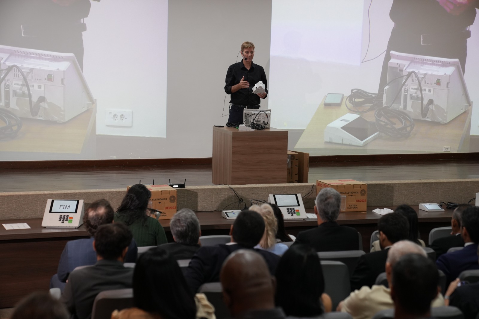 Durante apresentação técnica, um demonstrador exibe componentes internos de uma urna eletrônica sobre uma bancada, evidenciando o funcionamento do equipamento