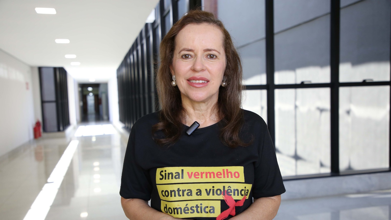 Juíza Cirlene de Assis posa para a câmera em um corredor do Fórum. Ela veste uma camiseta da campanha "Sinal Vermelho contra a Violência Doméstica", usa colar de pérolas e brincos claros. Ao fundo, aparecem cadeiras e portas do ambiente interno da unidade judiciária.