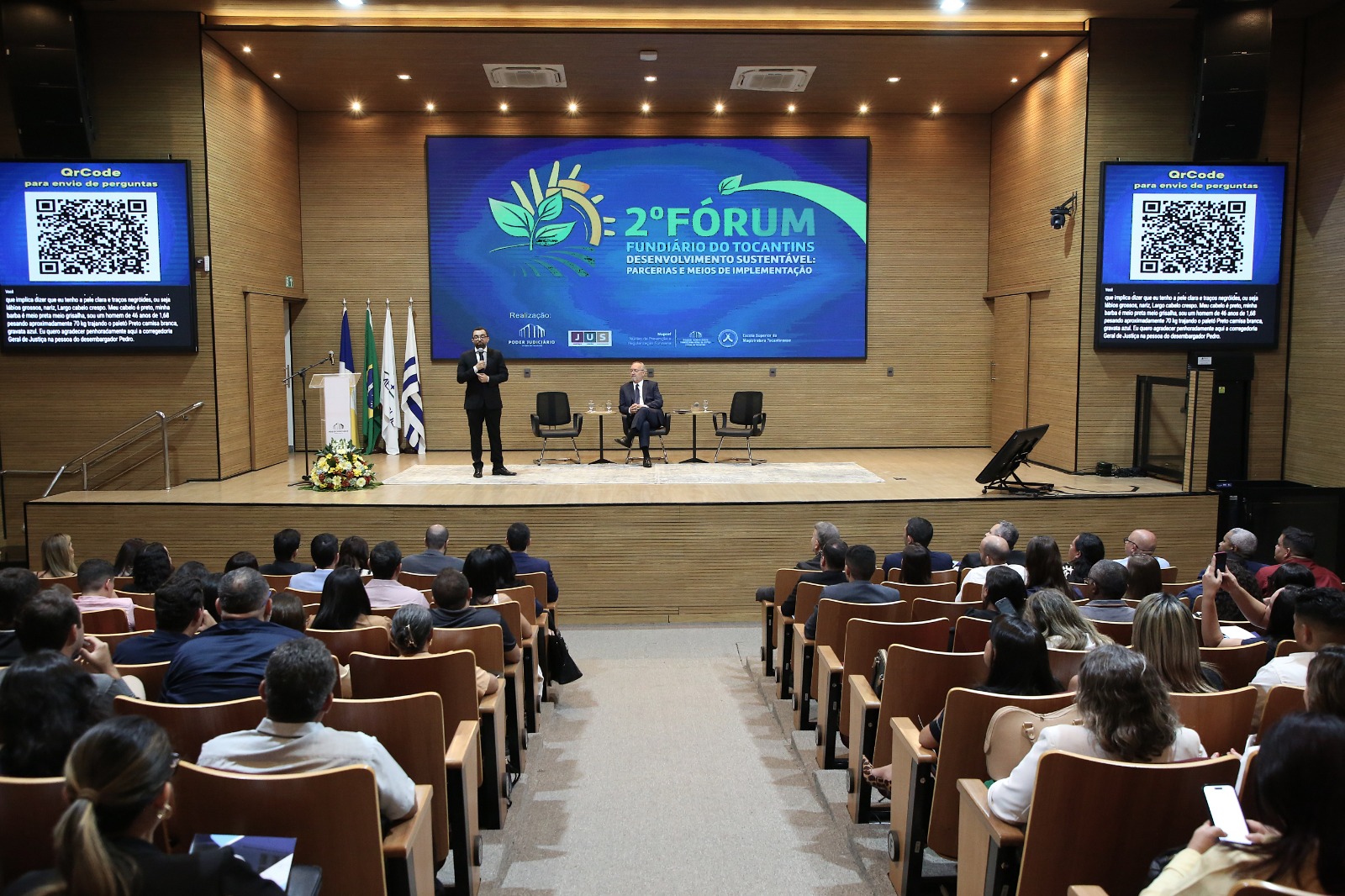 Fotografia em plano aberto do auditório da Escola Superior da Magistratura Tocantinense (Esmat), com público ocupando as poltronas e voltado para o palco. Ao centro, um palestrante em pé faz sua apresentação ao lado de outro participante sentado. No telão principal está projetada a identidade visual do 2º Fórum Fundiário do Tocantins, com o tema "Desenvolvimento Sustentável: Parcerias e Meios de Implementação". Nas laterais do palco, dois monitores exibem um QR Code para envio de perguntas. Bandeiras institucionais e um arranjo de flores completam o cenário.