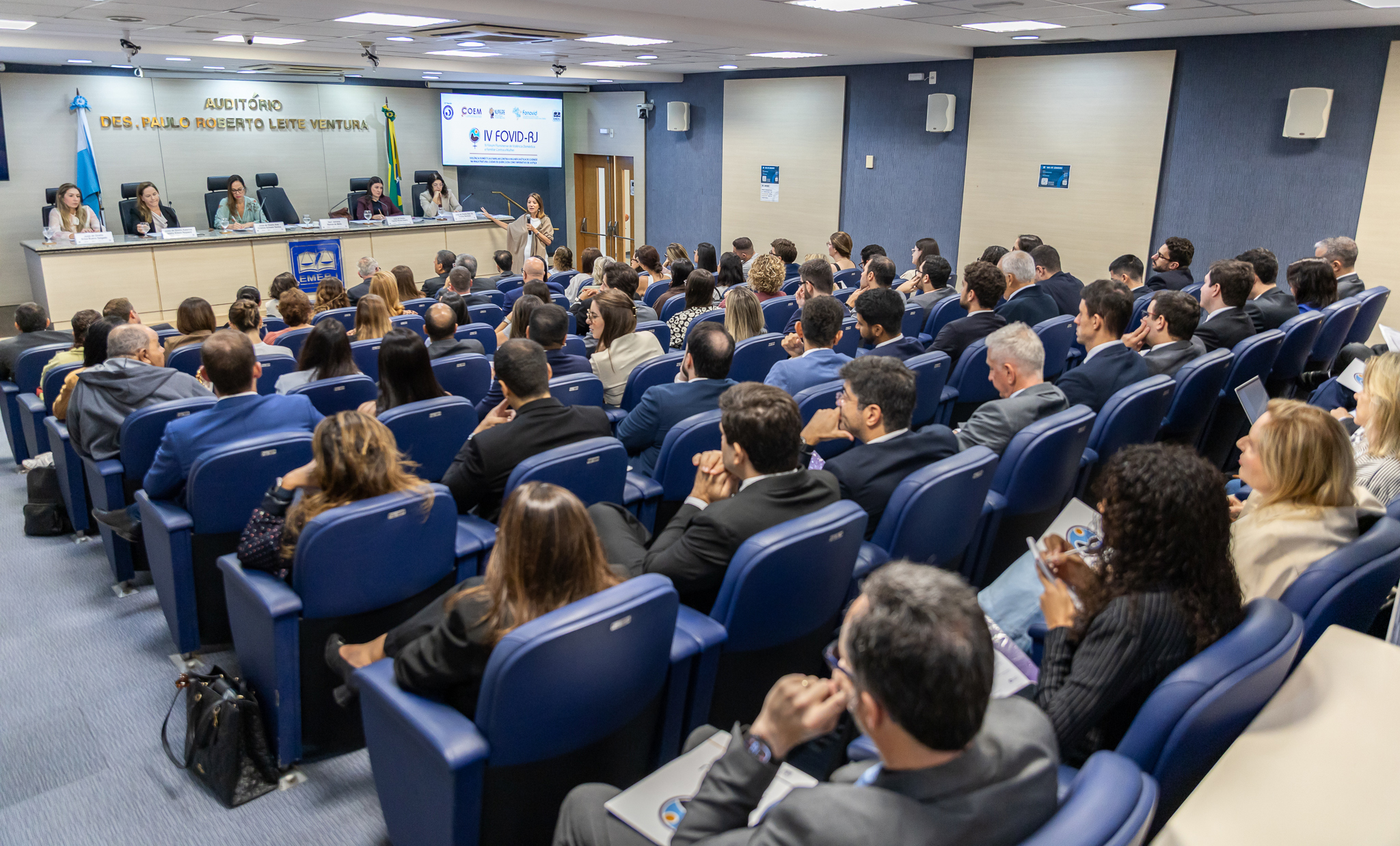 A imagem mostra participantes reunidos no auditório para acompanhar a programação do IV Fórum Fluminense de Violência Doméstica e Familiar contra a Mulher (IV FOVID-RJ). Ao fundo, uma palestrante se dirige ao público, enquanto integrantes da mesa acompanham a atividade.