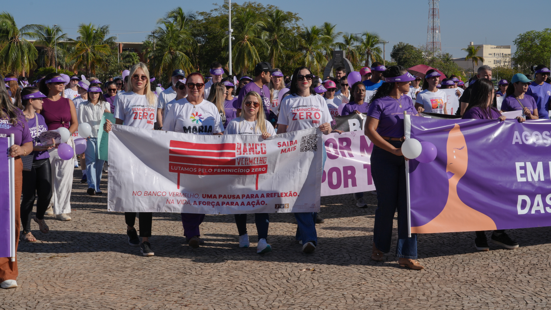 Imagem aberta da Caminhada do Agosto Lilás, com dezenas de participantes na Praça dos Girassóis, em Palmas. Em primeiro plano, mulheres seguram faixas de conscientização e combate à violência contra a mulher e ao feminicídio. Ao fundo, participantes vestem camisetas e acessórios nas cores branca e lilás.