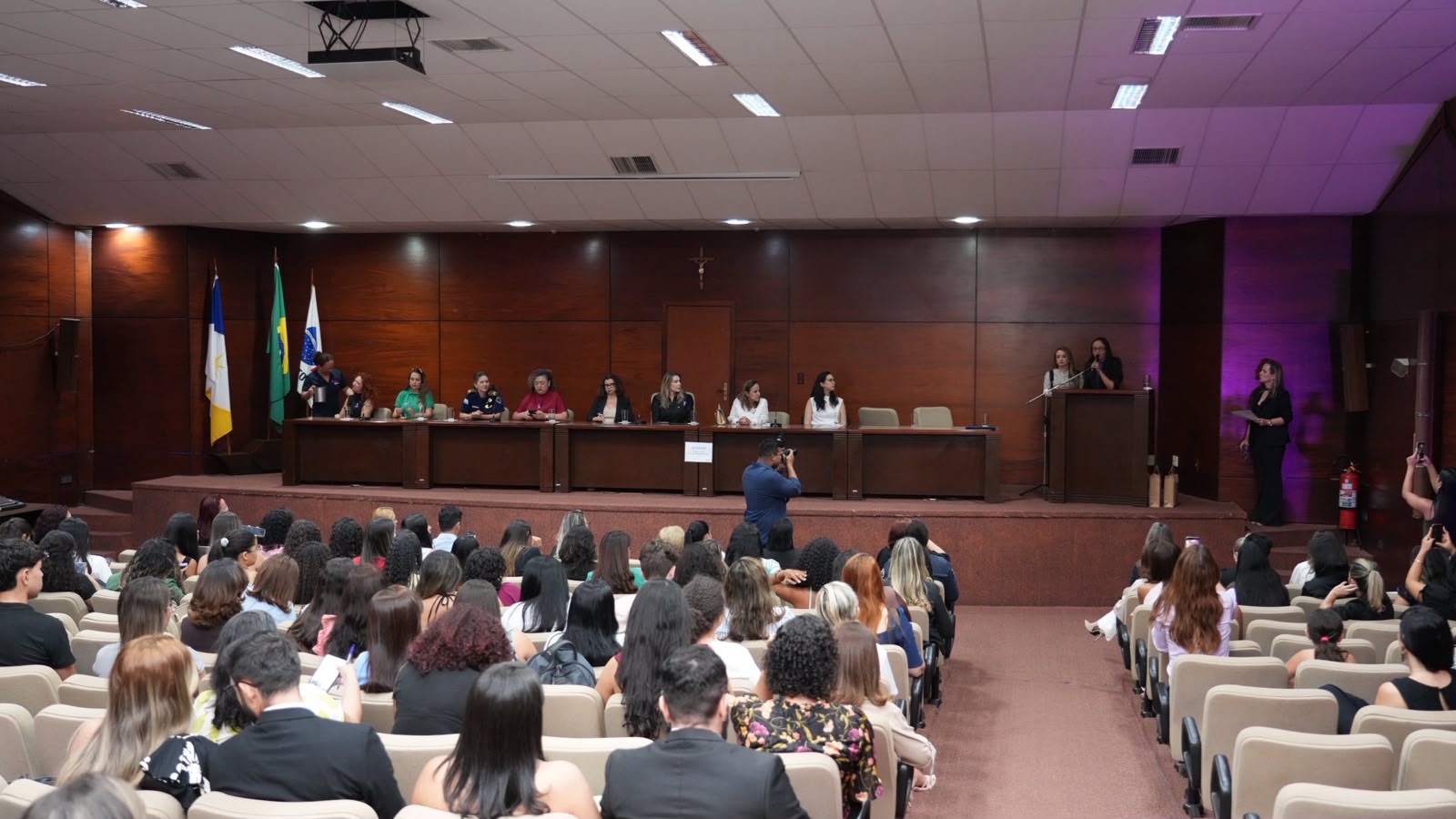 Imagem aberta do auditório da OAB onde foi realizado evento do Agosto Lilás; ao fundo a mesa com as autoridades e, em primeiro plano, o público sentado