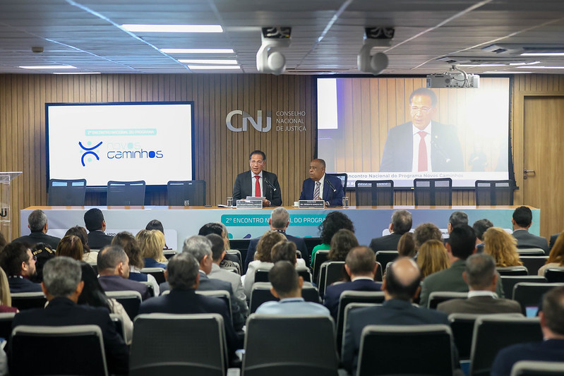 Fotografia mostra o auditório do Conselho Nacional de Justiça (CNJ) durante o 2º Encontro Nacional do Programa Novos Caminhos. À frente, dois representantes estão sentados à mesa principal, enquanto um deles faz uso da palavra. Ao fundo, um telão reproduz a imagem do palestrante e a identidade visual do programa. O auditório está lotado, com magistrados, servidores e demais participantes acompanhando a apresentação.
