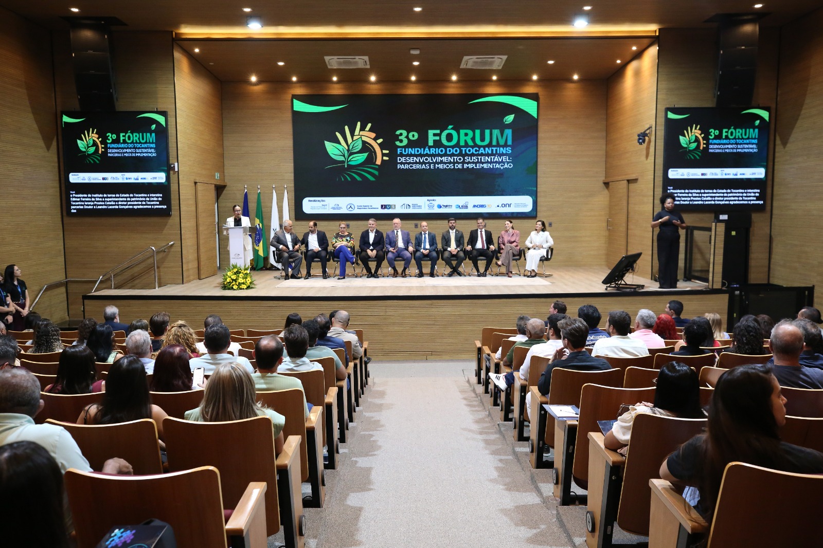 A imagem mostra o auditório do Tribunal de Justiça do Tocantins durante a abertura do 3º Fórum Fundiário do Tocantins, com o público acomodado nas poltronas e acompanhando a programação. No palco, há uma mesa composta por autoridades e representantes de instituições participantes do evento, além de uma pessoa na tribuna e uma intérprete de Libras.  Ao fundo, o telão principal exibe a identidade visual do 3º Fórum Fundiário do Tocantins, com o tema “Desenvolvimento sustentável: parcerias e meios de implementação”. As telas laterais reproduzem informações da programação, enquanto bandeiras institucionais e nacionais estão posicionadas ao lado do palco.