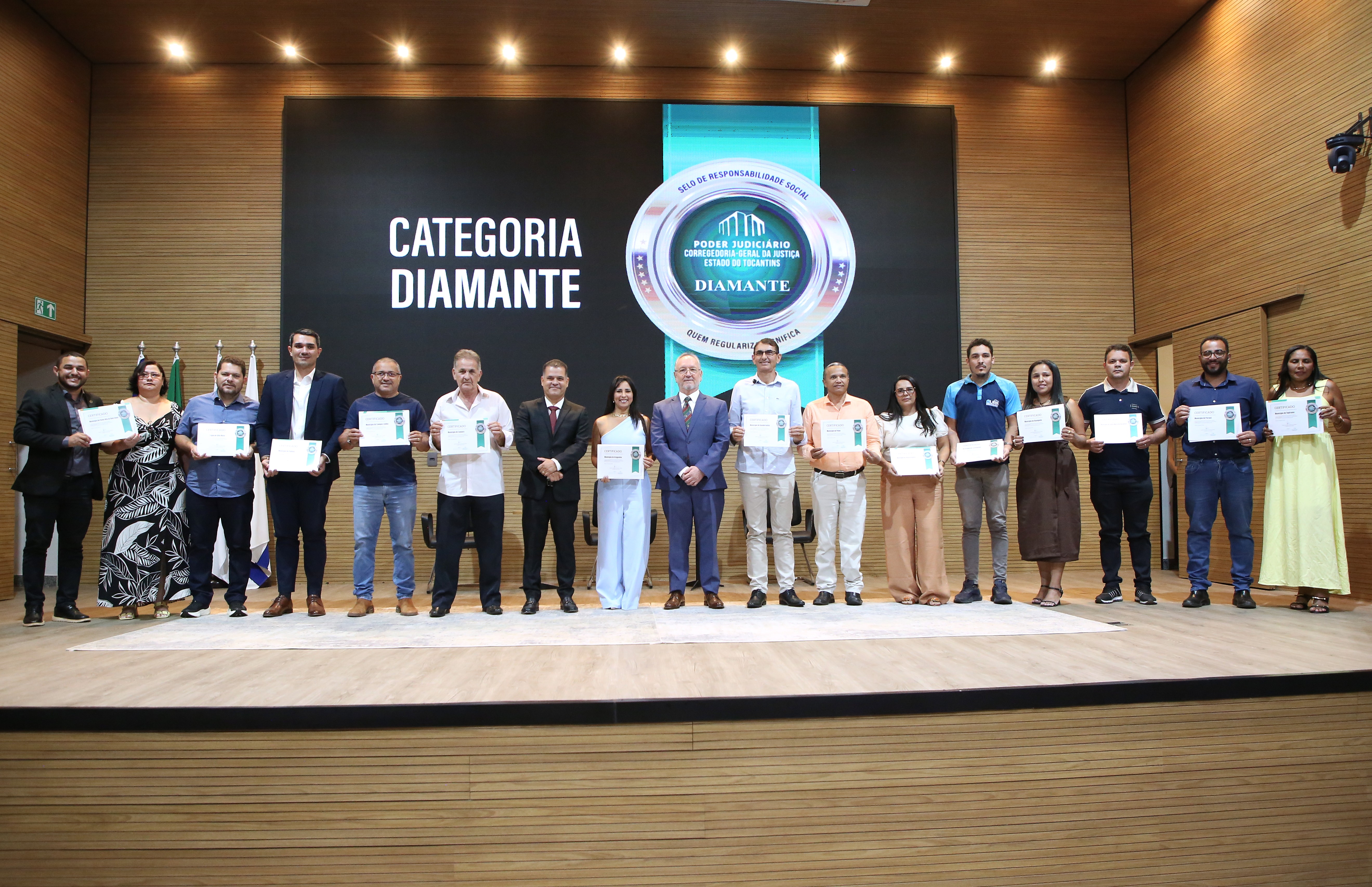 A imagem mostra várias pessoas em pé no palco do auditório do TJTO, segurando certificados durante o III Fórum Fundiário do Tocantins. Ao fundo, o telão exibe um selo na categoria Diamante.