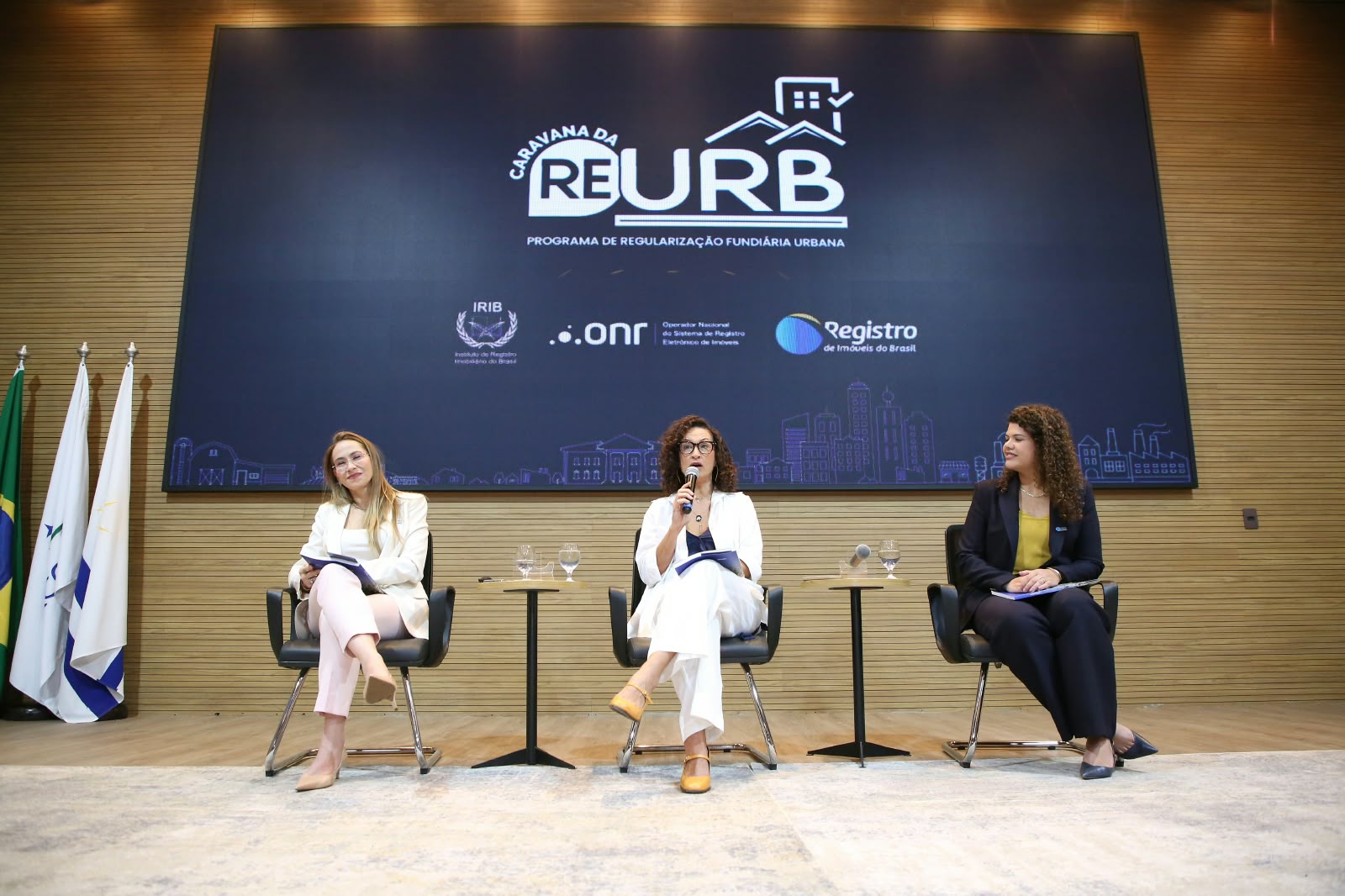 A imagem mostra três mulheres sentadas em poltronas no palco do auditório do TJTO, durante III Fórum Fundiário do Tocantins. Ao centro, uma das participantes segura um microfone e fala ao público, enquanto as outras duas acompanham a atividade. Ao fundo, um painel exibe a identidade visual da Caravana da Reurb – Programa de Regularização Fundiária Urbana, com as marcas das instituições parceiras. À esquerda, estão posicionadas as bandeiras institucionais.