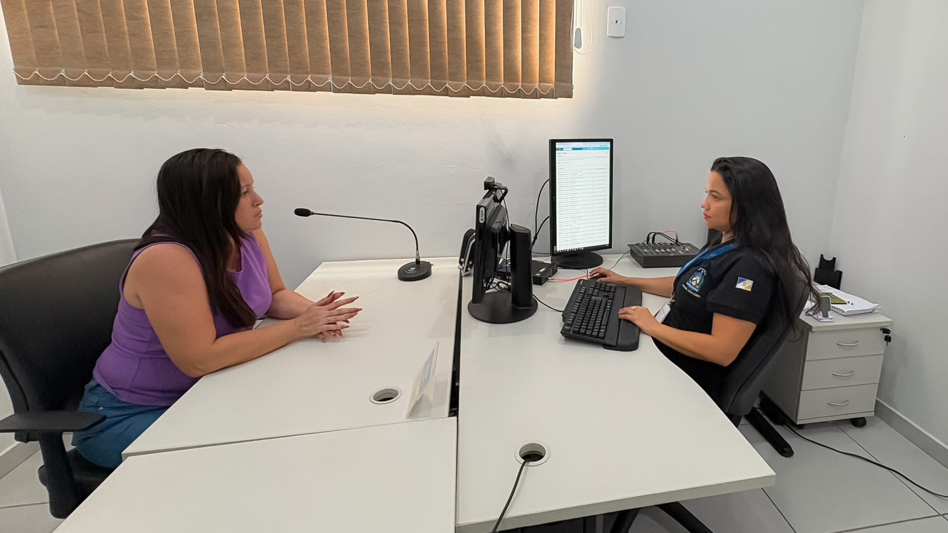 A imagem mostra duas mulheres sentadas frente a frente em uma sala de atendimento. Uma delas usa blusa roxa e a outra veste uniforme preto, diante de um computador. Sobre a mesa há microfones, teclado e equipamentos de informática.