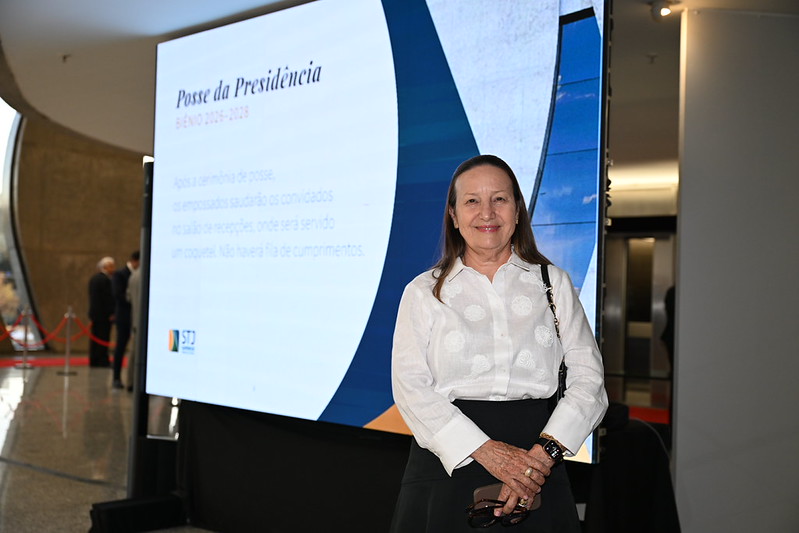 Presidente do TJTO, desembargadora Maysa prestigia posse. Ela usa saia preta e blusa branca e aparece sorrindo. Ao fundo um painel com o nome posse presidência.