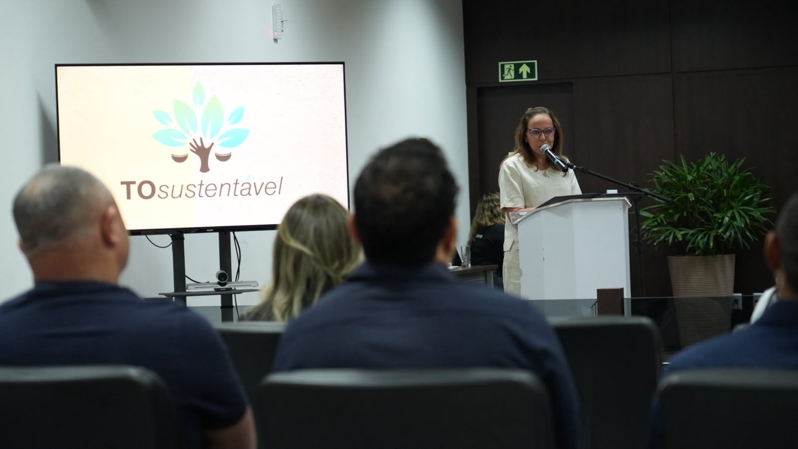 A imagem mostra a desembargadora Angela Prudente durante o lançamento do Projeto Coleta Seletiva Solidária do Tribunal de Justiça do Tocantins (TJTO), na Comarca de Miracema do Tocantins. A magistrada fala ao púlpito, enquanto outras pessoas aparecem de costas, acompanhando o momento. Ao fundo, uma televisão exibe o símbolo do programa Tô Sustentável.