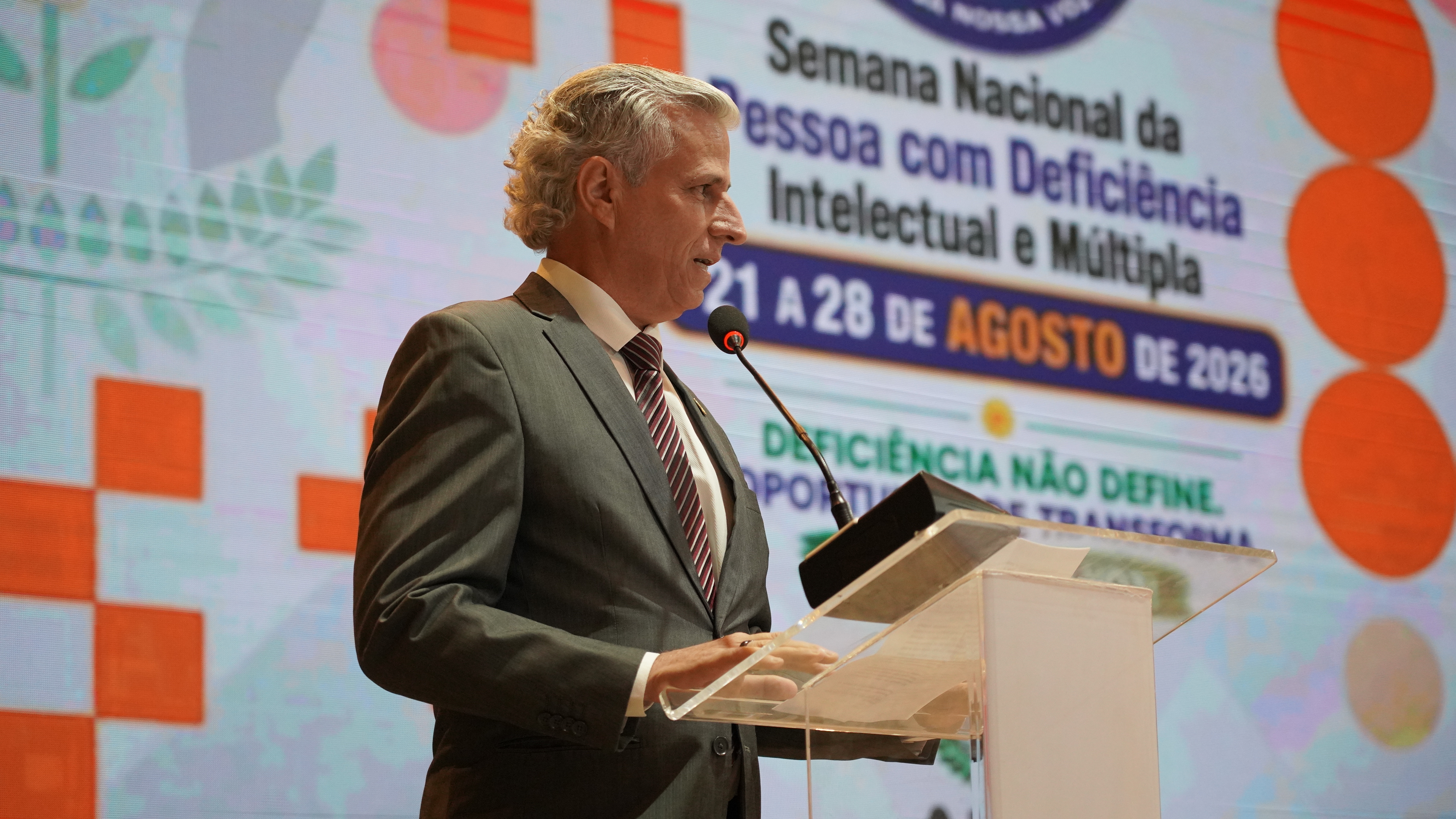 Juiz Esmar Custódio discursa durante a abertura da Semana Nacional da Pessoa com Deficiência Intelectual e Múltipla, realizada no auditório do Tribunal. Ao fundo, o painel apresenta o tema da campanha de 2026.