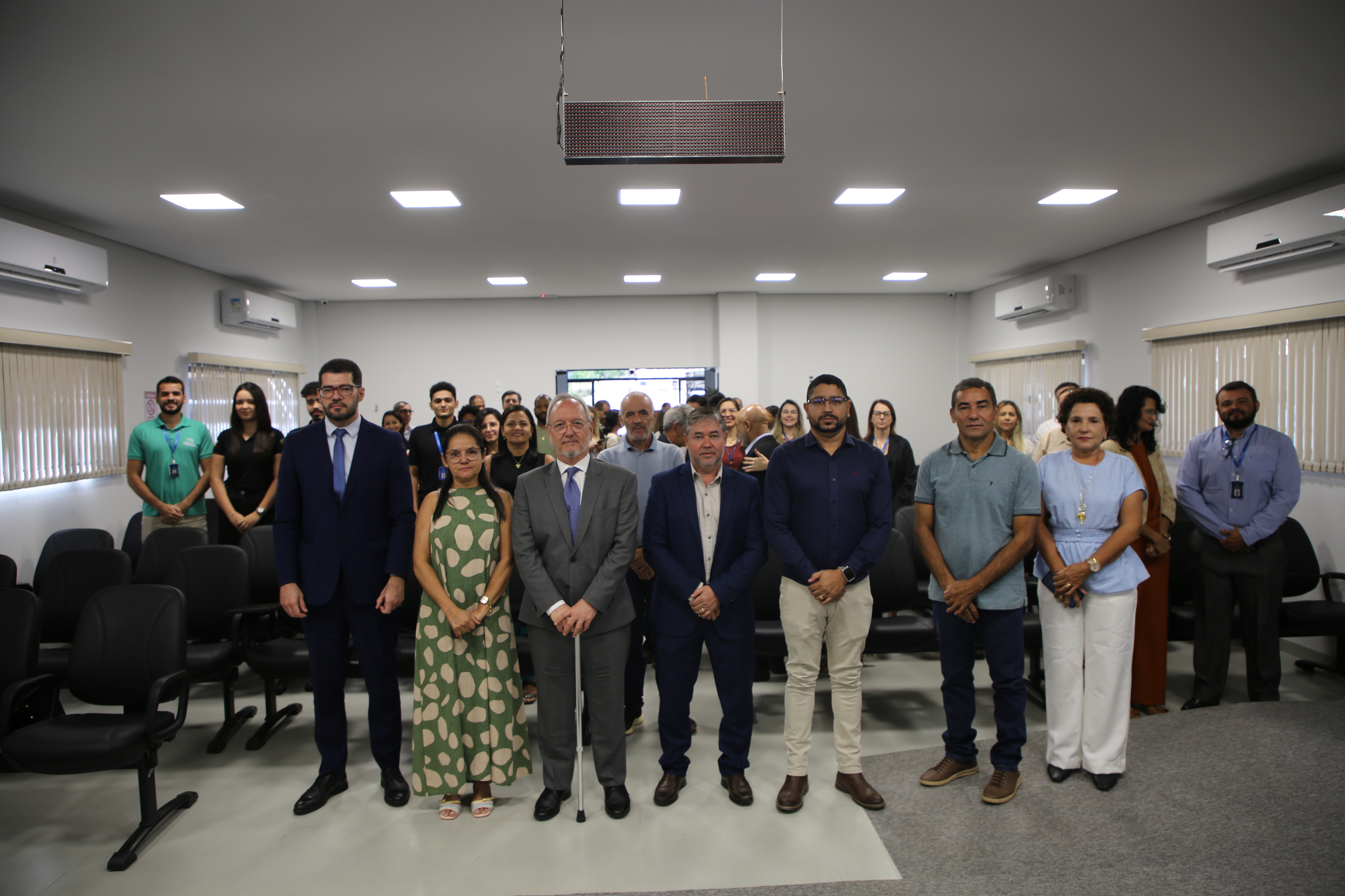 Grupo de pessoas posa para foto em um auditório. Na primeira fileira, sete pessoas estão lado a lado, enquanto outros participantes aparecem ao fundo, em pé entre as cadeiras. O ambiente possui fileiras de assentos, janelas laterais, aparelhos de ar-condicionado e iluminação no teto.