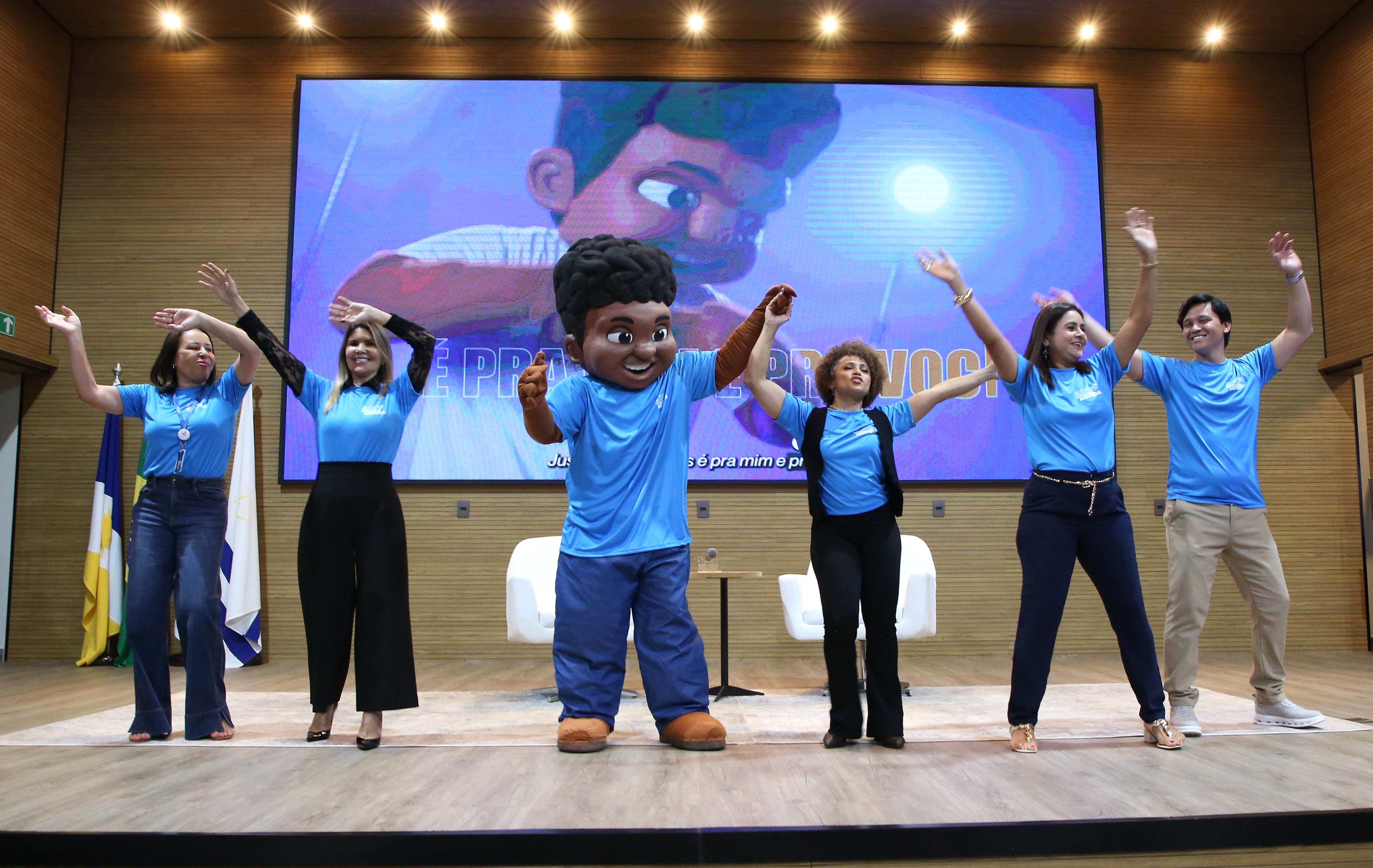 A imagem mostra o mascote Justino, do Tribunal de Justiça do Tocantins (TJTO), junto à equipe de fisioterapia do Centro de Saúde (Cesau), no palco do auditório do Tribunal, durante atividade de ginástica laboral na abertura da 11ª Semana da Saúde.